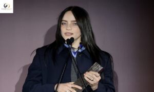 Woman speaking at a microphone during a public event, holding a small book or card in her hands.